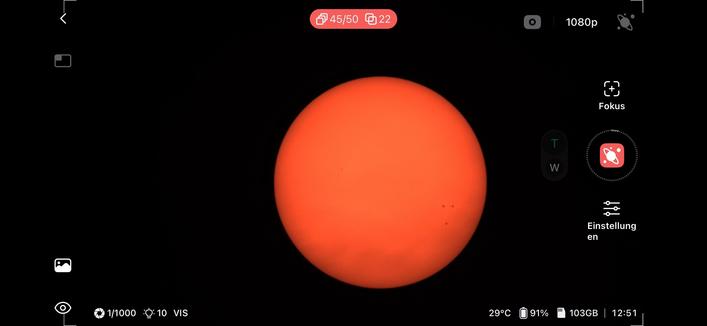 A bright orange sun is depicted against a black background. Small dark spots are visible on the sun's surface, suggesting solar activity. The image includes camera settings displayed in the corners.