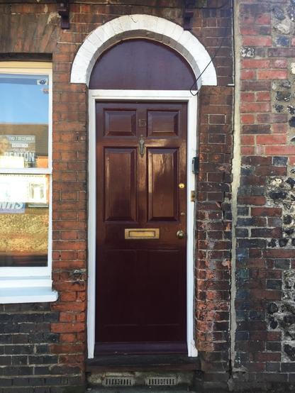 A maroon front door with an arch window above it. There is white trim around the door and it's set into a dark red brick wall. The number 15 is on the door above the knocker.