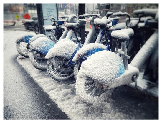 Photo of a row of blue CitiBikes parked at their station on a city sidewalk. The pavement is damp and the bicycles are covered in a thick layer of powdery snow.