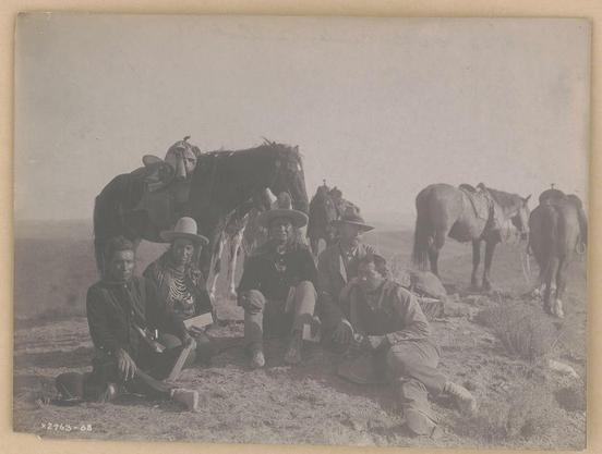 The image depicts a group of individuals and animals in an outdoor setting, likely during the late 19th or early 20th century. There are five people visible; four men seated on the ground forming a semi-circle with their horses behind them, and another individual sitting atop one of the horses.

One man is wearing traditional Native American attire, including a distinctive hat, while others wear Western-style clothing such as hats, jackets, and boots. They appear to be gathered around for rest or conversation during what seems like an expedition or journey across open land with sparse vegetation in sight.

Their expressions are relaxed; some look directly at the camera, suggesting they may have been aware of being photographed. The horses behind them wear saddles, indicating preparedness for travel on horseback, a mode commonly used by travelers and explorers during that era.