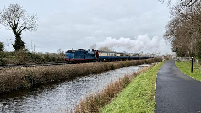 A landscape view of a blue steam locomotive running ‘backwards’ with it’s tender leading and smoke stack behind pulling a rake of vintage cream and blue carriages around a turn in the railway line and the canal running right next to it between the camera and the rails. The right of the frame is dominated by the paved towpath following the near-bank of the canal. A dull grey sky hangs overhead with  a cloud of clean white steam from the chimney contrasting nicely against it. Both edges are framed by bare trees.