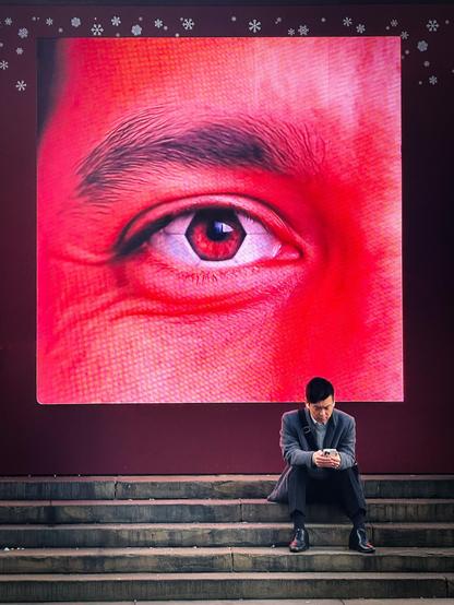 A man sits on the steps of the Eros statue at London's Piccadilly Circus looking at his phone. Above him on an LCD screen is part of a man's face shown in red, with a single large eye.