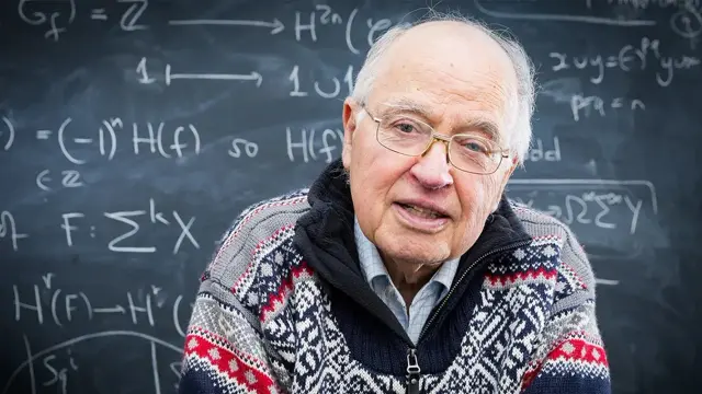 a person wearing a fair isle sweater seated in front of a blackboard wtih mathematical symbols
