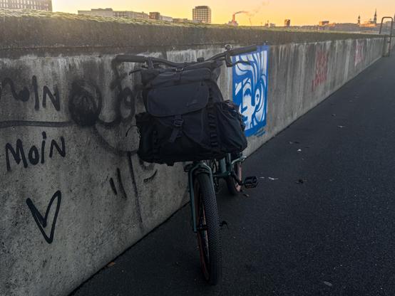 Das dunkelgrüne Brompton G Line von vorne an einer grauen Kaimauer mit blauweißen Graffiti. Vorne am Fahrrad das schwarze Lumabag Bromfort. Über der Kaimauer im gelblichen Sonnenuntergang die Skyline der HafenCity.