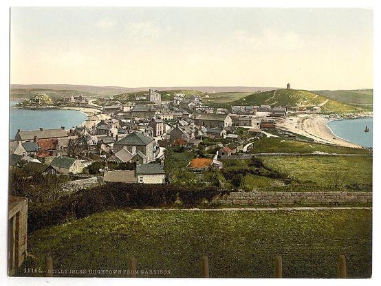 The image depicts a picturesque coastal town with an array of houses and buildings spread across the landscape, viewed from a high vantage point. The architecture varies in style but predominantly features gabled roofs and traditional construction materials such as stone or brickwork visible on many structures. A significant historical landmark stands prominently atop a hill to the right side of the scene, suggesting its importance within this setting.

A beach is discernible along the coastline with gentle waves lapping against it. The sea meets land in the distance where a harbor can be faintly observed, adding to the scenic beauty and hinting at local maritime activities. Greenery blankets much of the lower slopes leading up to the hilltop structure, indicating cultivated fields or perhaps pasturelands.

The overall coloration suggests an early 20th-century photograph with muted yet naturalistic tones; greens from vegetation, grays and browns on buildings, a turquoise sea, and pale blues in the sky. There is no visible modern infrastructure such as cars or contemporary clothing styles to provide precise dating but its historical context is implied by architectural design.

The image appears aged, possibly a century old given the photochrom process typically used during that era for color printing from black-and-white negatives. The upper left corner of the photograph c [...]