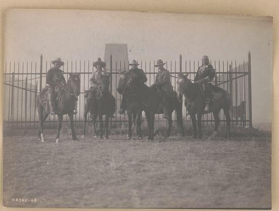 The image depicts a historical photograph of five individuals, presumably from the late 19th century. They are mounted on horses in an open field with dry grass beneath them and bare earth visible around their hooves. The subjects wear wide-brimmed hats, indicative of protection against sun or rain commonly associated with outdoor work environments.

Each person is adorned differently; some have decorative elements such as feathers or ornaments that signify status or role within a community context. They appear to be engaged in conversation but maintain an attentive posture toward their surroundings or each other.

The setting includes what seems like part of the Custer Monument, identified by its distinctive stone structure in the background with vertical lines and horizontal cross-bracing visible above it. The monument's purpose isn't explicitly clear from this perspective but suggests historical significance related to Native American history given context provided on the image caption which mentions "Custer Monument" and dates.

The overall color tone of the photograph is sepia, hinting at its age or artistic choice in presentation typical for that era’s photography. Notable features include detailed textures captured by early photographic techniques such as film grain, soft focus around moving elements like horses' legs indicating motion blur from a slower shutter sp [...]