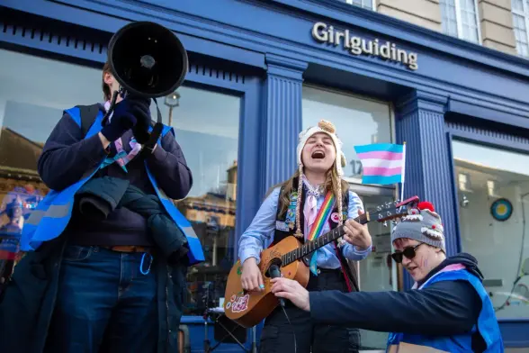A protestor holds a megaphone, while another plays guitar outside Girlguiding HQ.