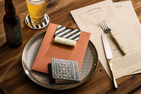 A stylish dining setup with a beer bottle, glass, menu, and utensils on a wooden table. Featuring a notable 'Townhouse Kitchen & Bar' design.