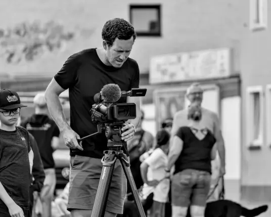 Ein Mann führt eine Kamera am Stativ. Ein Junge scheint ihm dabei zuzusehen. Die Kamera auf dem Stativ trägt ein großes Mikrofon mit Windschutz und einen kleinen Monitor, den der Mann konzentriert betrachtet. Um ihm herum bewegen sich viele Personen. Es ist nicht zu sehen, was die Kamera sieht. 
.
A man is holding a camera on a tripod. A boy seems to be watching him. The camera on the tripod has a large microphone with a windshield and a small monitor, which the man is looking at closely. There are lots of people moving around him. It is not possible to see what the camera is filming. 
.