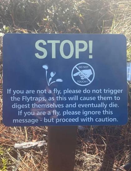A blue sign with the word "STOP!" at the top

Illustration of fly trap next to a "do not touch" symbol

"If you are not a fly, please do not trigger the Flytraps, as this will cause them to digest themselves and eventually die. If you are a fly, please ignore this message - but proceed with caution."