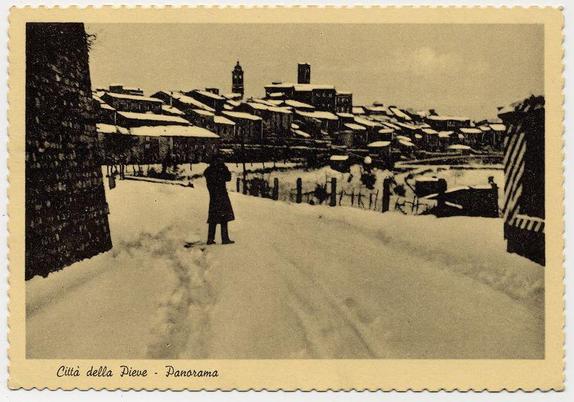 The image depicts a sepia-toned postcard of Città della Pieve, Italy during winter. A solitary figure is shown walking on snow-covered streets in the foreground with their back to the viewer. The town's architecture features traditional buildings with sloped roofs covered in snow, and there are visible tracks from foot traffic or vehicles left by a horse-drawn cart passing through. In the background stands a prominent clock tower that rises above the surrounding structures, indicating it may be an important local landmark. This postcard appears to capture a tranquil yet picturesque winter scene of this historic Italian town.