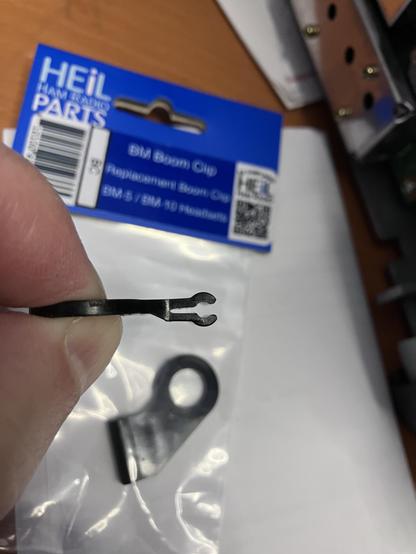 Black plastic clip that holds metal mic boom to earpiece as part of Heil BM-10 headset for my amateur radio station.  Package in background is a plastic bag with a bright blue cardboard tag that reads "HEiL HAM RADIO PARTS".