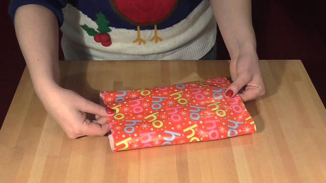 Hands demonstrating mathematical gift wrapping technique on rectangular present with colorful holiday pattern paper.