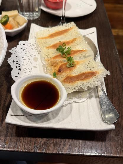 Plate with 5 pot stickers held together with a fried slurry. Front, small bowl of dumpling sauce. Right, large spoon. Back left, pickles.