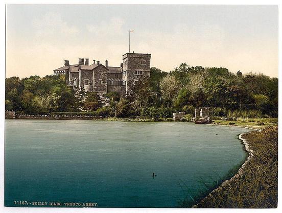The image depicts a historic stone mansion surrounded by lush greenery, situated beside a body of water. The architecture includes multiple chimneys and what appears to be a flagpole with a small red flag at the top. There is also an inscription indicating that this location is Scilly Isles on Tresco Abbey in Cornwall, England, between approximately 1890 and 1900.