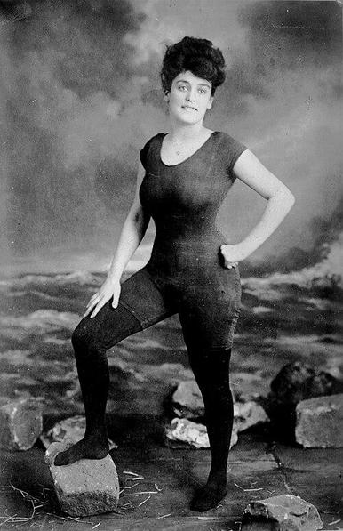 Kellermann in her famous one-piece bathing suit, c. 1906. She is fair-skinned with dark hair styled in an updo. Her facial expression is a slight, confident smile. She is wearing a dark, form-fitting swimsuit that covers her from her shoulders to her ankles. The swimsuit appears to be made of a textured material, possibly wool or a similar fabric. Her stance is confident and poised, with one leg bent and placed on a stone, and her hand on her hip. Her other arm is slightly bent, with her hand resting at her side.

The background is a painted backdrop depicting a seascape. The artist has created a cloudy sky with rough seas, providing a backdrop for the model. Scattered around her are some stones, adding to the illusion of a shoreline setting. The lighting appears to be soft and diffused, typical of studio photography from the period, creating a somewhat ethereal quality. The overall composition is a classic studio portrait, designed to showcase the subject's form and confidence.