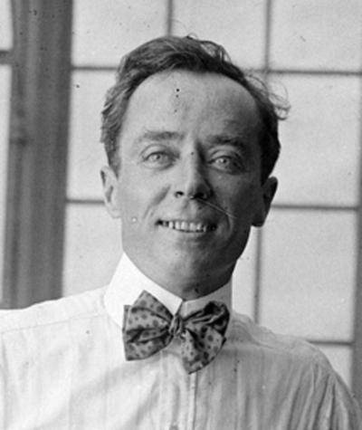 Herbert Brenon, 1916. 

He is positioned in the center of the frame, from the chest up. The man has short, dark hair that is slightly receding. His eyes are light-colored and he has a slight smile, revealing his teeth. He is wearing a white collared shirt with vertical stripes and a patterned bow tie. The bow tie has a mottled pattern of dark and light spots. The background appears to be a window with a grid pattern, though the details are blurred. The lighting is soft and diffused, creating subtle shadows on his face. The overall composition is simple and classic, with the focus entirely on the man's expression and attire. The image has a vintage feel, likely due to the black and white format and the style of the man's clothing and hairstyle. There are some minor imperfections like dust spots on the image surface, which are common in older photographs.