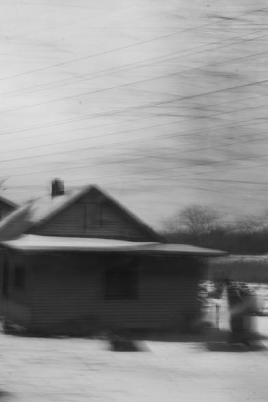 Black and white photo of a country home. The tones are mostly underexposed and shadow-ey. Even the sky is a fairly mid gray, and the snowy front yard maybe barely passes the 60% lightness mark. Across the sky, there is an interesting cross-hatching pattern of faint lines. The ones going diagonally up-and-right are electrical wires. The ones going the other diagonal are phantom pixels from foreground trees that blurred almost to invisibility because the car was moving as this shot was taken out the passenger window. The house is similarly blurred, but in ways that aren't uniform and leave certain parts captured at different offsets/times. The overall effect is like a house being swept away by the early stages of a tornado in a dream.
