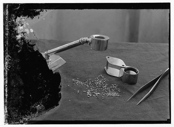The image is a black and white photograph featuring various tools commonly used in the diamond industry, such as magnifying glasses, tweezers, and trays. The background consists of scattered diamonds or small stones on what appears to be a tablecloth. This photo captures items from Jewish factories involved in diamond production during 1939 March at Plain of Sharon & along the coast to Haifa Tel Aviv Diamond Works where cut and polished diamonds are ready for sale according to additional information provided.