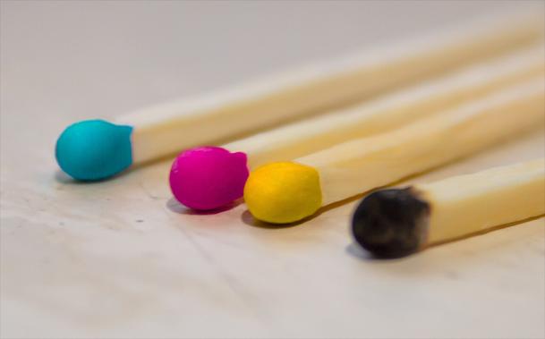 A photo of four matches, one cyan one magenta one yellow and one black. As in fire-lighting matches, with different colour heads, closeup, sitting on a table.