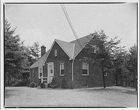 The image depicts a two-story brick house with several notable features, including multiple windows and a chimney on the roof. The structure has white trim around its doors and window frames, contrasting against the dark bricks of the exterior walls. A concrete porch can be seen at ground level in front of what appears to be an entrance door. Surrounding this residence are tall trees indicative of a wooded setting or neighborhood with ample greenery. This monochrome photograph might have been taken between approximately 1920 and 1950, as suggested by the accompanying text information about George Moss houses. The photographer's name is Horydczak, whose works span around that time period (approximately 1890-1971).