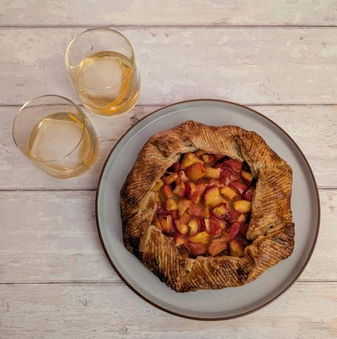 1) A square photo of a peach galette and two cocktails sitting on a white wooden background. The galette has a golden-brown crust decorated with diagonal lines, filled with a shiny peach filling that is a mixture of reds, pinks, oranges and yellows. The cocktails are whisky-coloured drinks in low-ball glasses with a giant ice cube each and an orange peel garnish.
