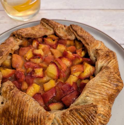2) A close-up square photo of the same peach galette and a cocktail sitting on a white wooden background. The camera is angled so that you are looking right across the peach filling, and can see big chunks of ripe peach in a shiny syrup.