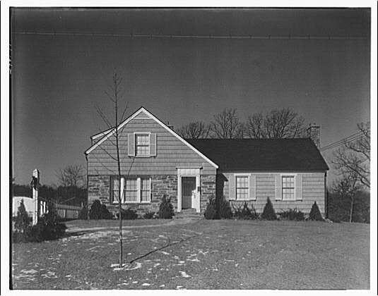 The image depicts a two-story residential house with a mix of brick and siding exterior walls, located in what appears to be a suburban neighborhood. The house features several windows, including one on the front facade that has shutters, and multiple smaller window units along its side elevation. There is a small tree growing near the foundation of the home, and another larger tree stands behind it.
In the foreground, there's an empty yard with some patches of grass visible and what looks like a few trees or shrubs planted around. The sky above appears clear, indicating that this photo was taken on a sunny day without any significant cloud cover. Overall, the setting seems calm and peaceful, likely in late winter given the presence of snow.
This black-and-white photograph provides an insight into residential architecture from approximately 1920 to 1950, as indicated by its inclusion in George Moss houses documentation. The source suggests that this particular house is located at or near 114 Pierce Drive and was captured around a century ago during the early part of the twentieth-century architectural style transition.
The image has been credited to Horydczak, an individual active approximately between 1890-1971. The photograph appears on the website for George Moss houses with its specific source URL provided in the description.