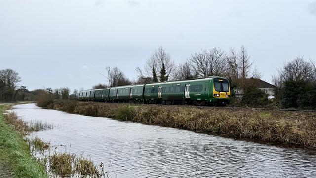A landscape shot of a dark green railcar-type train with white doors and a yellow front approaching from left to right along the far bank of a choppy canal with a dull grey sky overhead and a line of bare trees behind.