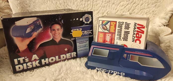 Auf einem weißen Kunstfell liegt links eine Schachtel und rechts ein Diskettenhalter. Der Diskettenhalter entspricht in der Grundform dem aus ST:TNG bekannten Shuttle und ist Dunkelblau mit aufgeklebten Fenstern/Verzierungen. Er ist oben offen. Darin stehen mehrere CD. Die Vorderste ist eine Leser-CD des Mac Magazins. Die Schachtel zeigt einen Jungen in einer roten ST:TNG Kapitäns-Uniform der den Diskettenhalter, gefüllt mit 3,5 Zoll Disketten hochhält.