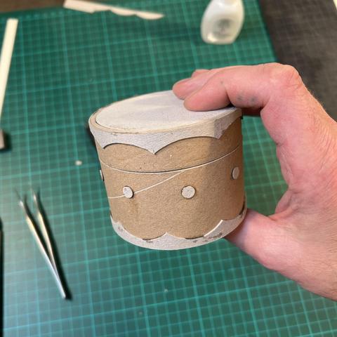 My hand holding what looks a bit like a brown and grey cardboard drum - upon closer inspection it turns out to be a small, cylindrical box, decorated with an embossed wavy border at the top and bottom, and a row of dots around the middle