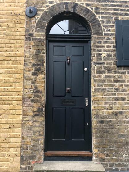 A sleek black door with a knocker shaped like a hand, and a smart semicircular fan light above it. Yellowish brick is around the door and a number 16 has been subtly painted on the door.