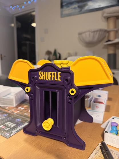 A purple and yellow 3d printed device with the word "shuffle" on the front. The boardgame "wingspan" is in the background.
