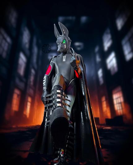 A low down POV shot looking upwards to a neoprene clad Jackal, welcoming you to 'inspect' his boots. One foot is raised off the ground, almost as if implying there's more than just an inspection.

He is inside an abandoned building, backlit with the bright moonlight and an orange light lower down. His shiny body and cape are reflecting the light. His bright green eye stands out amongst the contrasting colours.