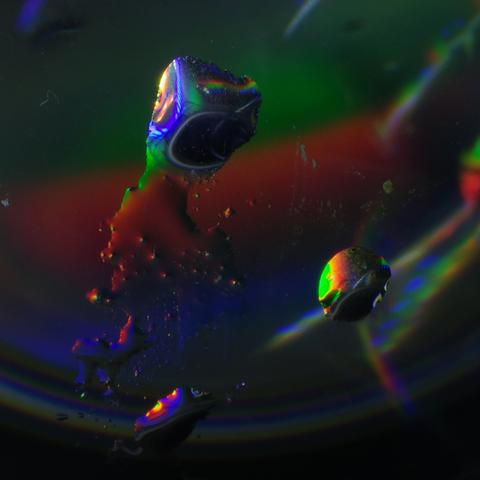 Four irregular water drops on a large glass sphere dominate the image. The largest resembles an angry octopus screaming at two of the drops, its rage-distorted mouth resulting from the reflection of a hole in a CD, appearing so black because the photograph was taken at night, along with two narrowed eyes and furrowed brows. The drops, also heavily distorted, reflect another CD, both of which are leaning against the sphere but are not visible in the frame. The sphere, of which only a portion is visible in the photograph, rests on a CD, also out of frame. The CD surfaces refract the white LED light illuminating them into their spectral colours.