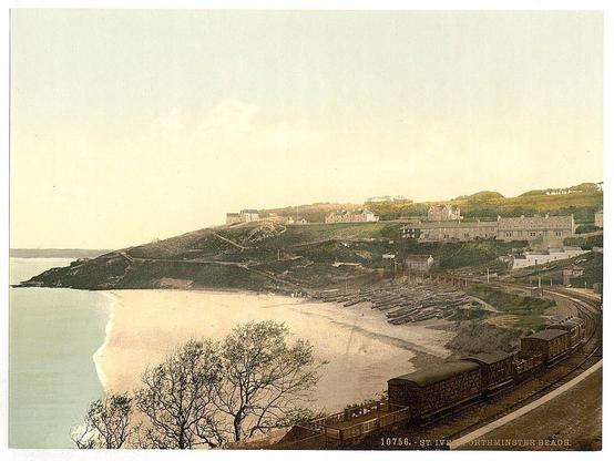 The image depicts a picturesque coastal scene from St. Ives, Porthminster Beach in Cornwall, England during the late 19th or early 20th century. The panoramic view shows a sandy beach extending into the distance where it meets calm sea waters on one side and gentle hills with greenery on the other.

On the left edge of the image, there is a cliff leading down to the water's edge, which gradually slopes downward towards the central part of the picture where the beach spreads out. To the right, we can observe rows of wooden boats laid horizontally in shallow waters adjacent to what appears to be a boatyard or storage area.

The background features an array of buildings indicative of either residential areas or possibly hotels and facilities catering to visitors at Porthminster Beach. These structures are distributed along the top edge of the image on elevated ground, with some nestled into the hillsides suggesting that they were built for scenic views over the coastline.

A railway line is visible in the foreground running parallel to the beachfront. Alongside it, we can see freight wagons carrying various types of goods or materials, hinting at active transportation and commerce during this period.

The overall atmosphere exudes a sense of tranquility with no apparent human activity captured within these boundaries. The color palette consists mainly of muted greens from th [...]