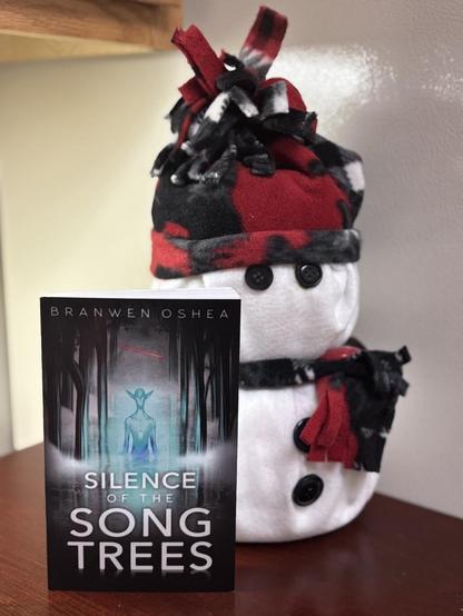 A paperback of Silence of the Song Trees sits on a table next to a plush snowman. The cover of the book features a cerulean-colored alien with long pointed ears standing in water surrounded by sad-looking trees. A mist hovers above the water. A red streak of light passes over the alien’s head.