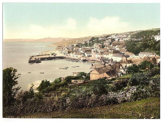 The image depicts a picturesque coastal town with densely packed buildings situated along the shoreline. The architecture of the houses suggests an older, possibly historic settlement, characterized by steeply pitched roofs and varied rooflines. Many structures have chimneys visible against their facades, indicating residential use. A few larger boats are moored in calm waters near the harbor, which extends into a bay or inlet with additional small vessels scattered throughout.
The town is set on hilly terrain leading down to the water's edge, where rocky outcrops protrude from the sea. The landscape includes verdant vegetation and trees that partially obscure certain buildings, adding depth and texture to the scene. A stone wall runs alongside a grassy slope in the foreground, providing a natural barrier between the viewer and the town.
In the background, rolling hills rise up against a clear sky with soft clouds, hinting at a peaceful day or early evening ambiance. The overall color palette is muted yet warm, featuring shades of green from foliage, earth tones for the buildings, and various hues in the water reflecting sunlight. This image likely captures an everyday scene of life within this coastal town during its heyday as depicted by St. Mawes' close proximity to Falmouth, Cornwall.