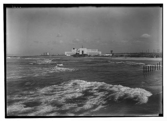 The image depicts a black and white photograph of an industrial facility situated by the sea. The building appears large with multiple columns, possibly indicative of its significance or function as a power house or processing plant. In front of this structure is turbulent ocean water with visible waves crashing against what seems to be breakwater pillars emerging from the sea. Additionally, several tall poles extend into the sky parallel to the shoreline, which could serve for various purposes like navigation markers or electrical lines.
The overall atmosphere appears somber and industrialized due to its grayscale coloration and the overcast skies above. The text in the photo indicates it is an American Colony (Jerusalem) photograph from 1934-1939 by G-Eric & Edith Matson, possibly linked with a collection at Tel Aviv's Reading Power House or related institutions.
Additional information suggests that this image might be part of a larger set held in the collections associated with The Reading Power House.