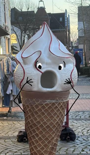 Eine große Eistüten-Statue mit einem überraschten Gesicht und karikaturhaften Armen ist in einem öffentlichen Bereich ausgestellt. Der Hintergrund zeigt Geschäfte und Straßenelemente mit einem Kopfsteinpflaster.