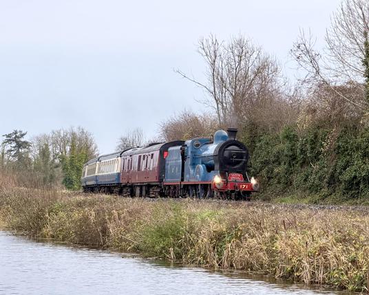 A telephoto front and side view of a blue steam locomotive with a black front and red running boards pulling a Burgundy brake van and a rake of cream and blue vintage carriages. There is no steam coming from the chimney, but there is a hint of a heat haze as the engine coats. A red name board on the front of the locomotive reads “Santa Special”.  The line is on the far bank of a canal which is visible in the foreground. There is a line of bare trees behind.