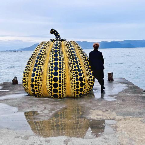 Ein Kunstwerk am Meer: Ein großer gelber Kürbis mit vielen schwarzen Punkten unterschiedlicher Größe, auf einem nassen Anleger, der ins Meer führt. 
An der rechten Seite steht eine rothaarige Frau, kleiner als der Kürbis. Sie ist schwarz gekleidet, und lehnt sich zum Meer gewandt an den Kürbis.