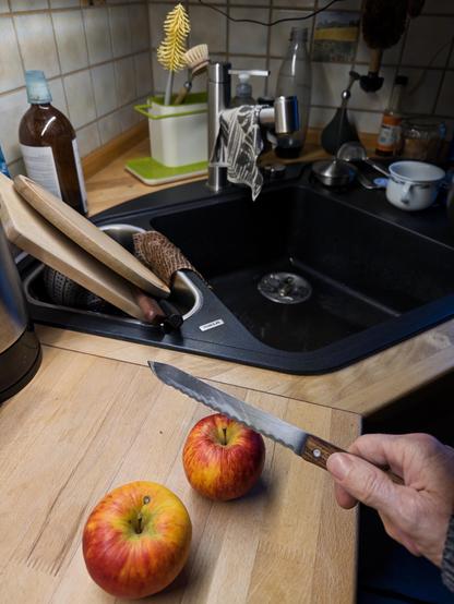 Eine Küchenszene mit zwei roten und gelben Äpfeln auf einer Holzarbeitsplatte. Eine Hand mit einem Messer befindet sich in der Nähe eines der Äpfel. Im Hintergrund sind Küchenutensilien, ein Spülbecken mit Geschirr und verschiedene Flaschen zu sehen.