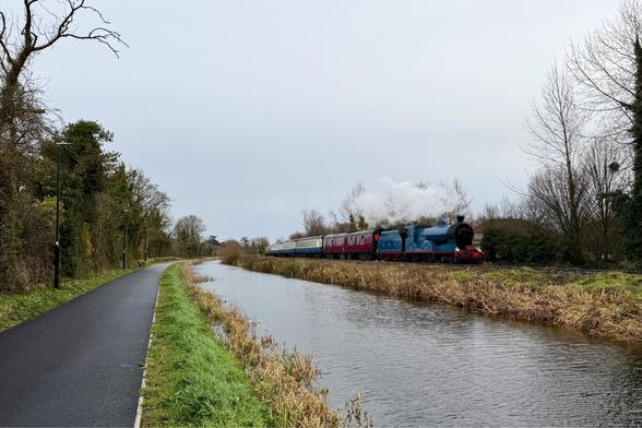 A landscape view looking down the paved towpath of rural canal with a blue steam train approaching along the far bank. The path, canal, railway, and a pair of bare hedge lines framing both ends of the scene run perfectly parallel from the right foreground towards a vanishing point in the left background. There is a small cloud of clean white steam from the steam locomotive which is pilling a burgundy brake van and a rake of cream and blue vintage carriages. The sky above is overcast and greyb