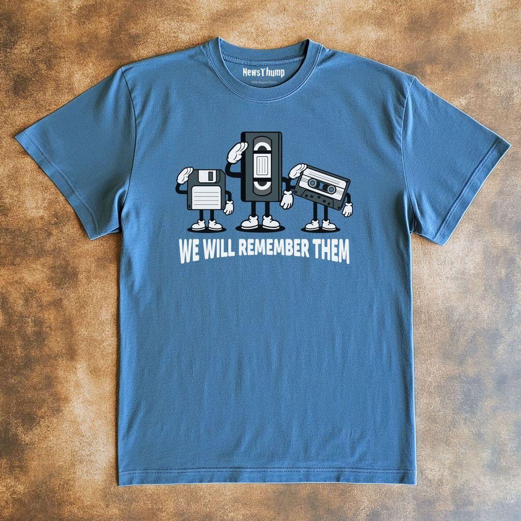 a blue t-shirt lying flat on a wooden surface.  On the front is a graphic design of a 3.5 inch floppy disc, a VHS cassette tape, and a C90 audio tape, saluting forwards.  Underneath is the text "We will remember them".