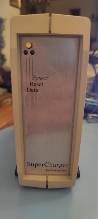 Ein schlankes beiges Gehäuse mit silberner Front steht hochkant im Bild. Am oberen Rand sind ein Taster und zwei Leuchtdioden im Dreieck angeordnet, der Taster oben. Von allen drei gehen Linien nach unten zur jeweiligen Beschriftung Data, Reset (fur den Taster) und Power. Am unteren Ende der Frontplatte steht in groß "SuperCharger" und klein darunter "by beta systems".
