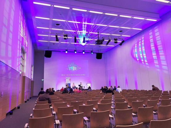 vortragsraum mit stühlen in violettem licht mit zuhörern sowie personen auf der bühne