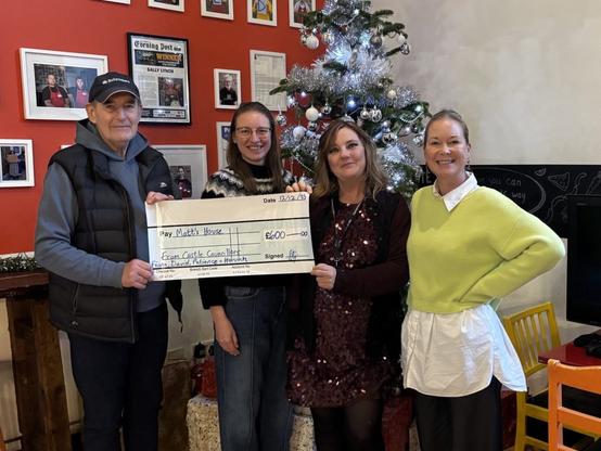 Four people standing indoors in front of a decorated Christmas tree, holding a large ceremonial cheque for £600. The setting includes festive decorations, framed photos, and a chalkboard wall with drawings and text.