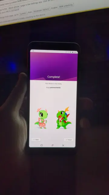 Фотография уже полностью фокусируется на телефоне с линуксом, на нём я чуть продвинулась по приветствующему экрану и на нём можно видеть двух талисманов КДЕ, динозаврика в розовом платье и динозаврика с прикольным красным шарфом.