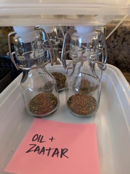 Image shows four small swing top bottles with zaatar spices in the bottom, and a label designating them as olive oil to be infused with zaatar.